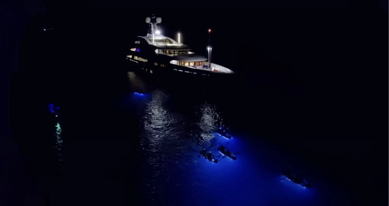 Illuminated clear kayaks rafted beside a brightly lit yacht at night