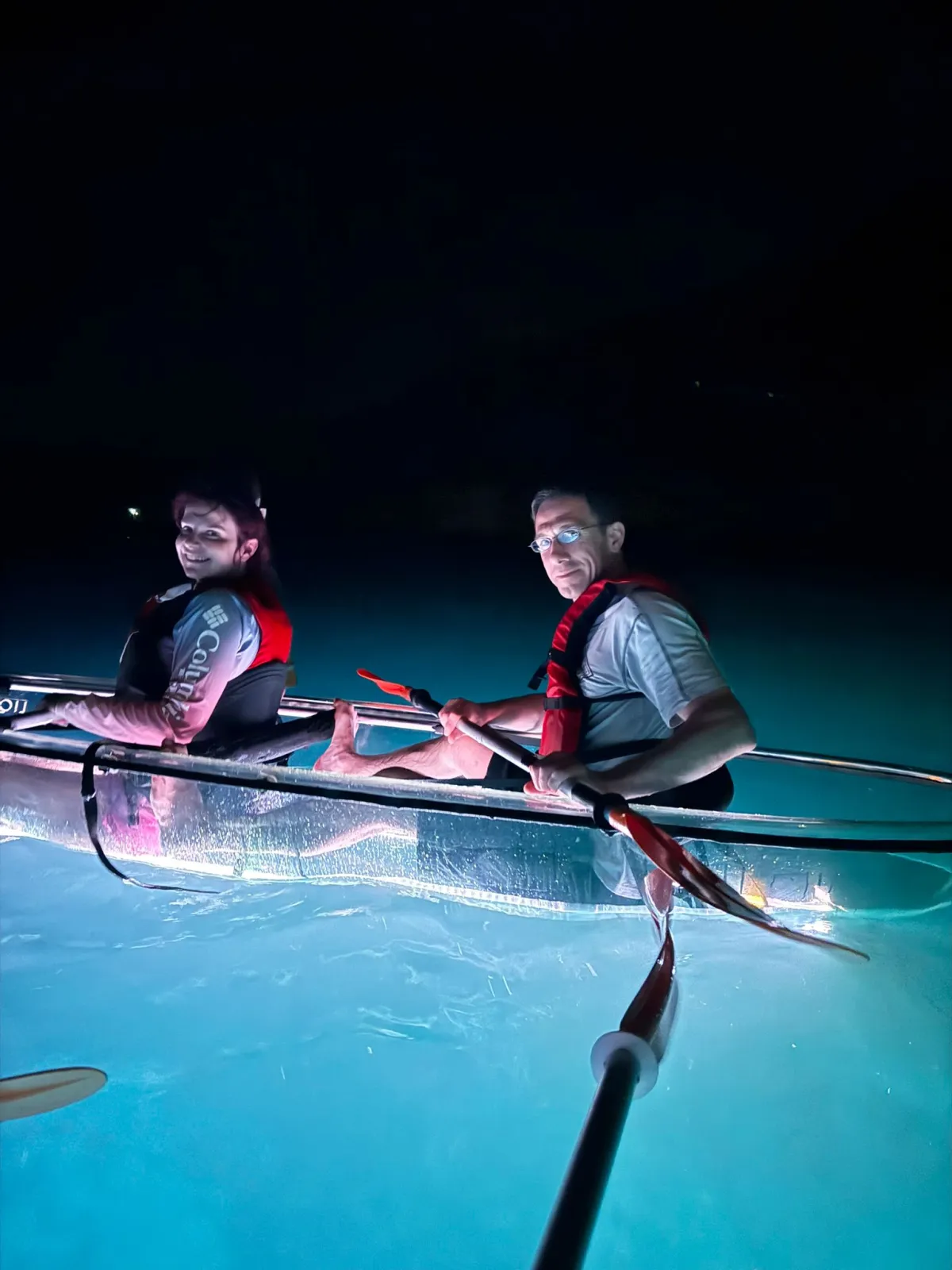 A couple smiling from an illuminated clear kayak on the dark bay