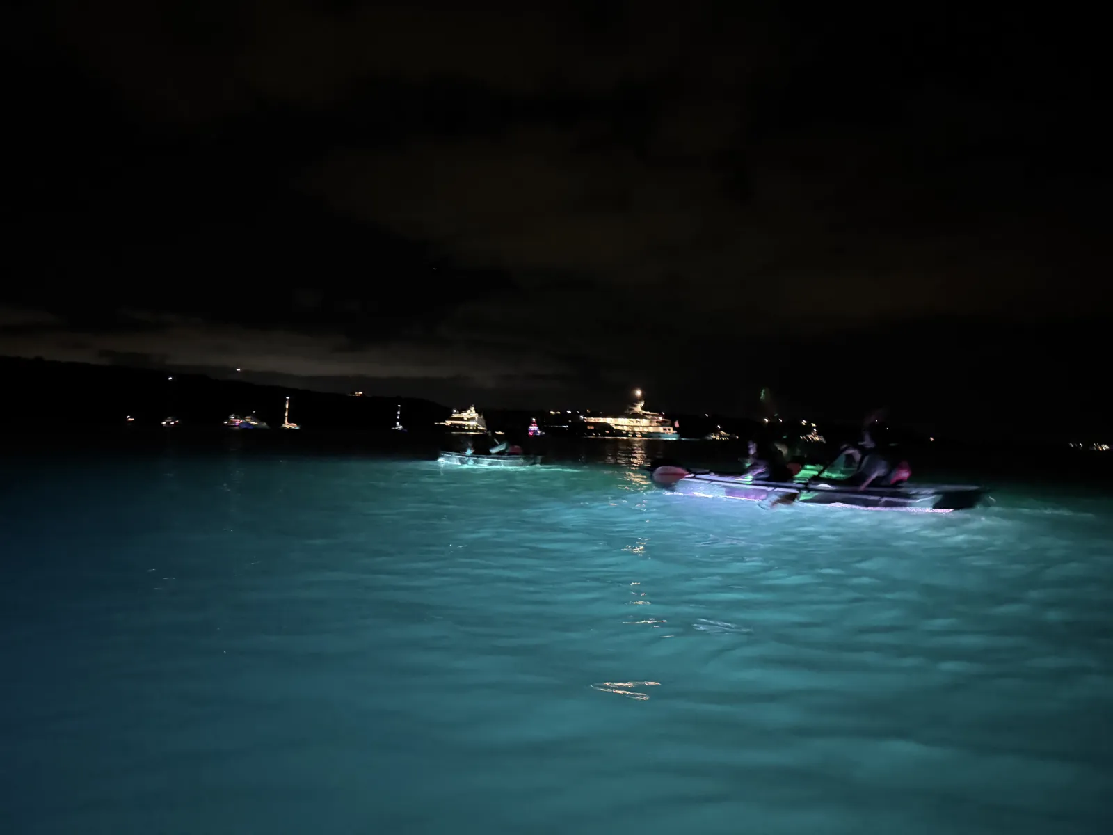 Lit clear kayaks on the open bay with anchored yachts glowing on the horizon