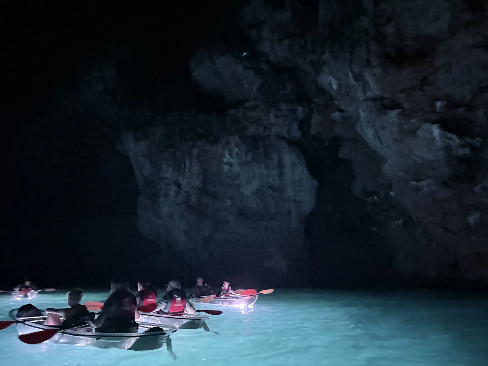 Paddlers in lit clear kayaks beneath a dark limestone overhang