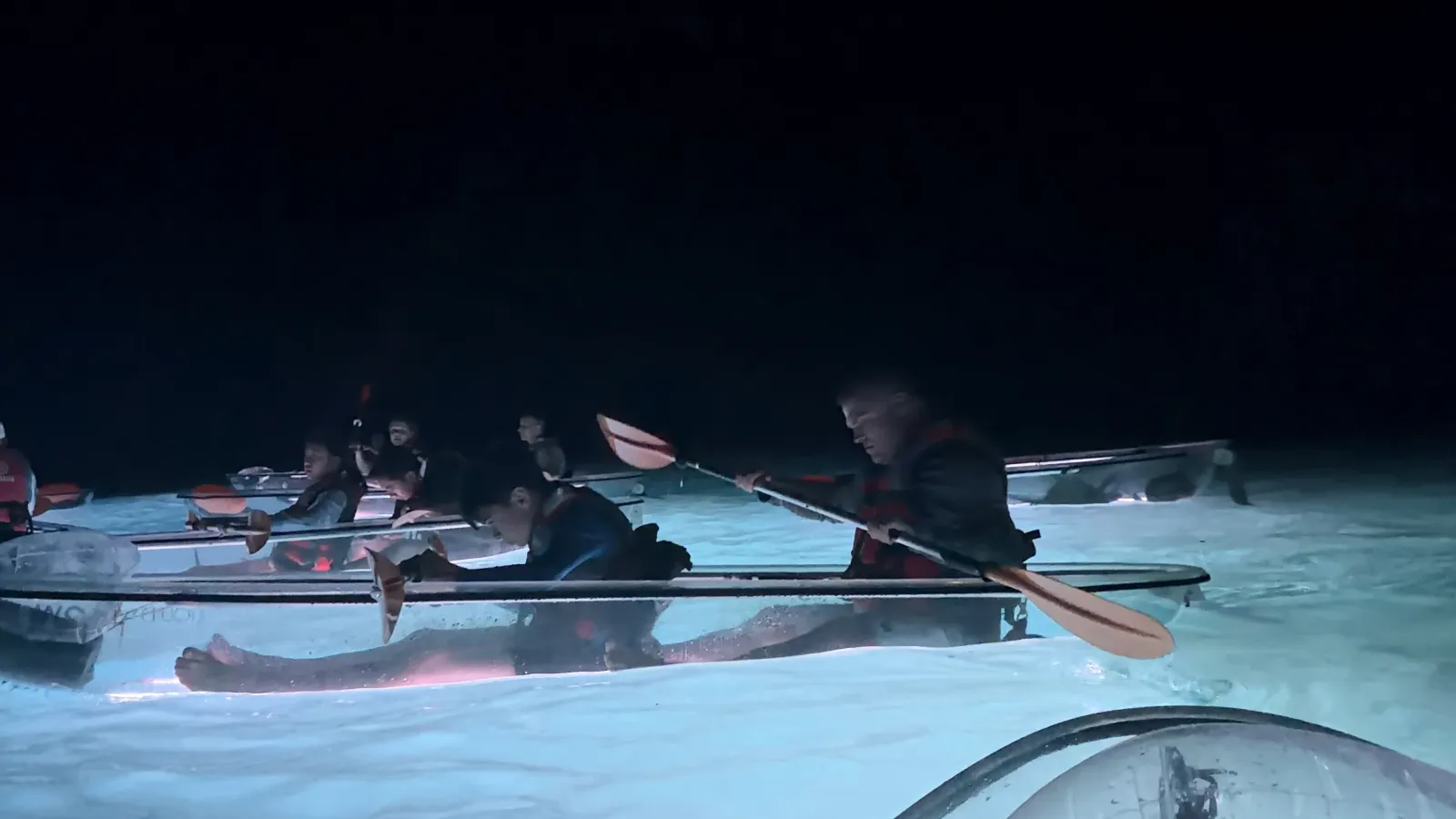 Paddlers up close, the LED hulls of their clear kayaks glowing white on the dark water