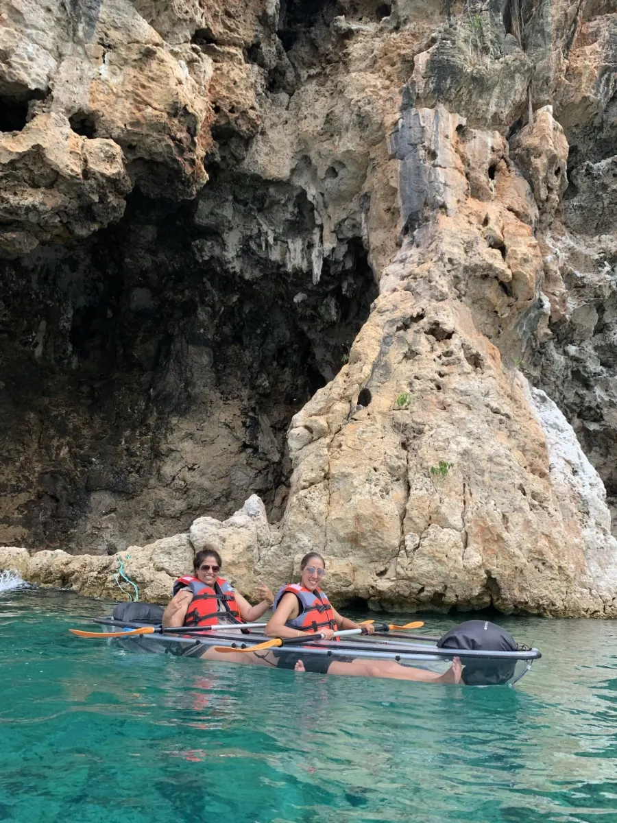 Two guests in a clear kayak by the sea caves, one throwing a shaka